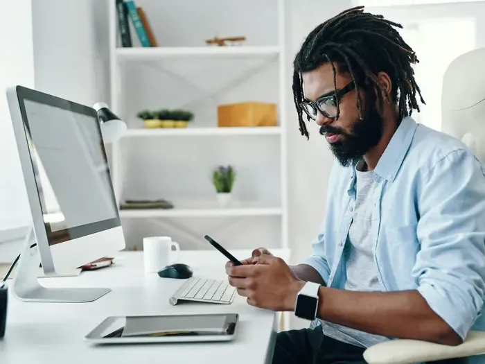 Entrepreneur wearing glasses uses a smartphone at a clean desk with computer, tablet and coffee in a bright modern office